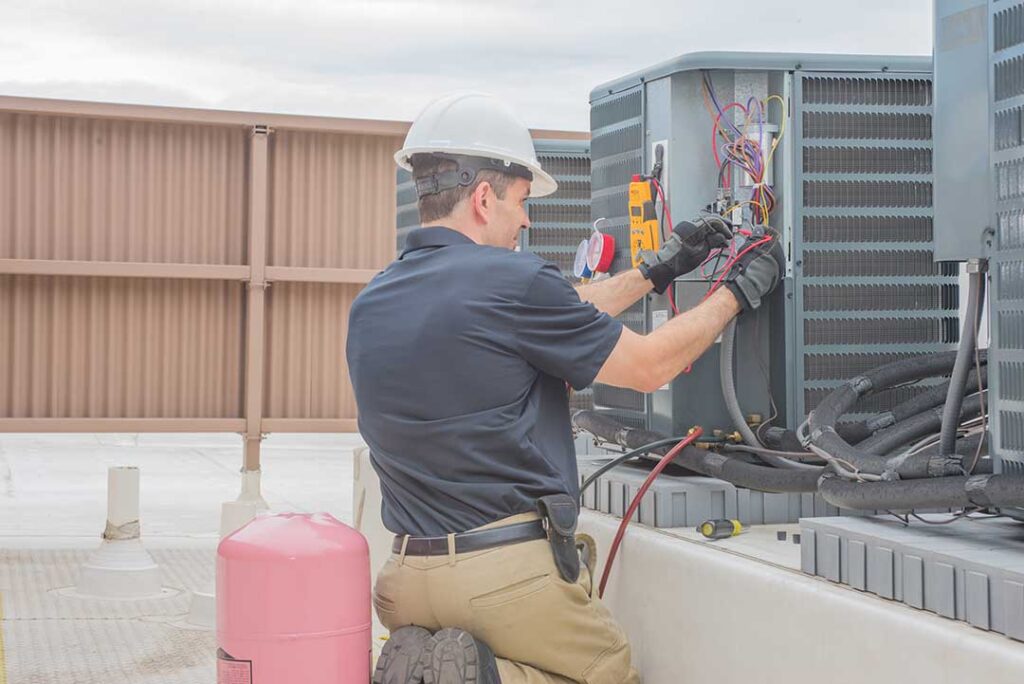 Homeowner gently rinses an outdoor AC condenser coil with a garden hose while wearing gloves.