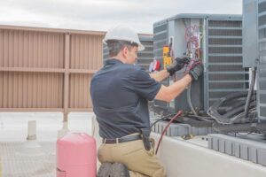Homeowner gently rinses an outdoor AC condenser coil with a garden hose while wearing gloves.