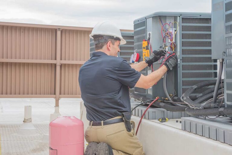 Homeowner gently rinses an outdoor AC condenser coil with a garden hose while wearing gloves.